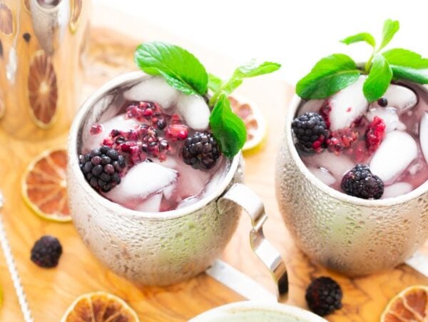 Two silver mugs with ice, blackberries, mint, and a vibrant purple Blackberry Moscow mule sit on a wooden board with dried citrus slices—an inviting cocktail with vodka that's perfect for anyone learning how to make a Moscow mule.