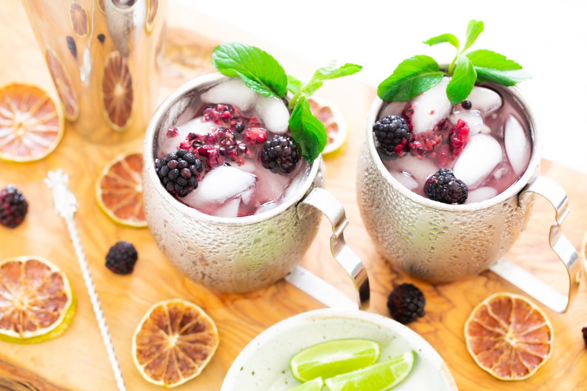 Two silver mugs of berry cocktails with ice, mint, and blackberries sit surrounded by dried citrus and lime wedges—perfect for trying a refreshing Blackberry Moscow mule at home.