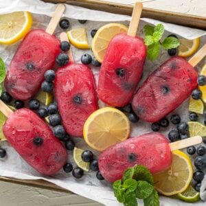 Six Blueberry Lemonade Popsicles on a tray with lemon slices, blueberries, lime slices, and mint leaves—a refreshing summer treat bursting with citrus and blueberry flavours.