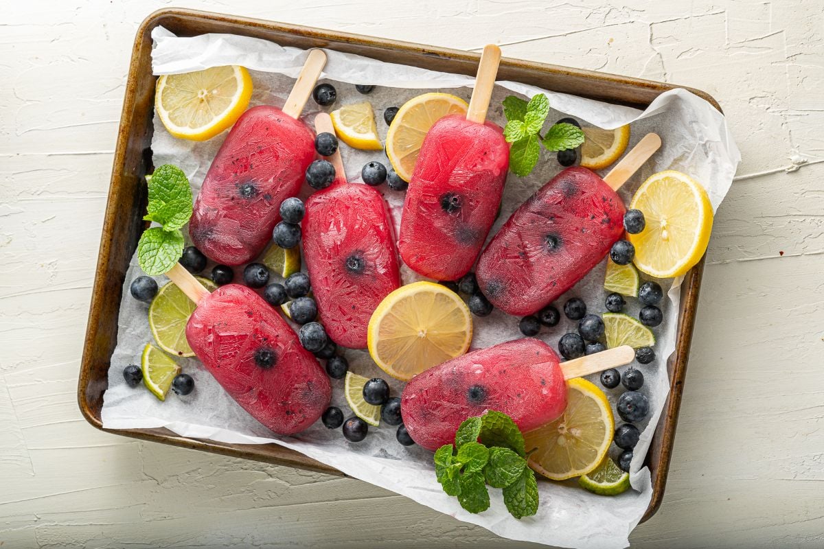 Six red citrus and blueberry flavoured popsicles with blueberries, lemon slices, and mint leaves on a tray lined with parchment paper.