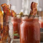 A mason jar of Bloody Mary with Clamato juice, garnished with bacon and olives, sits front and center, with more jars and festive garnishes in the background—a perfect cocktail for Canada Day.