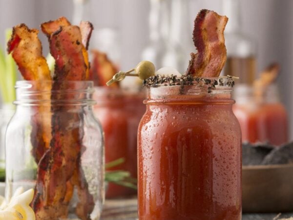 A mason jar of Bloody Mary with Clamato juice, garnished with bacon and olives, sits front and center, with more jars and festive garnishes in the background—a perfect cocktail for Canada Day.