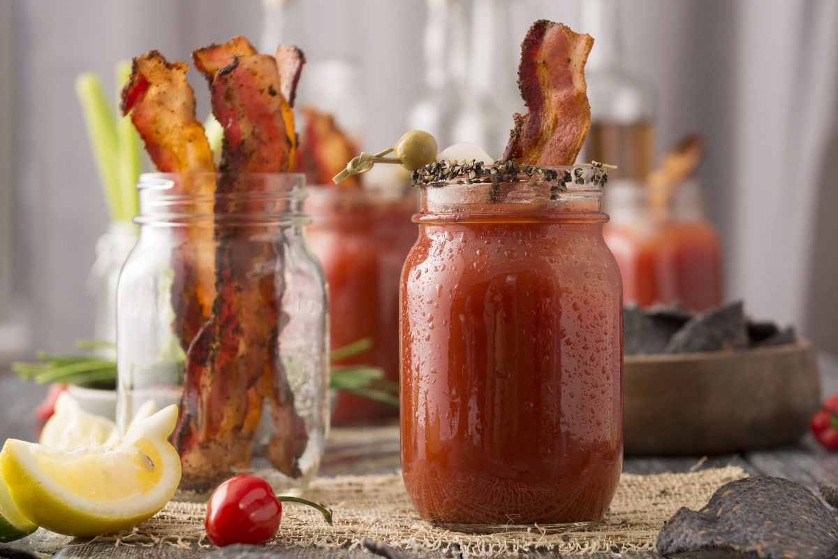 A Canadian Caesar Cocktail in a jar, garnished with bacon, olive, and pepper, with more bacon in a jar behind it—showcasing classic Caesar cocktail ingredients from Canada’s National Drink.