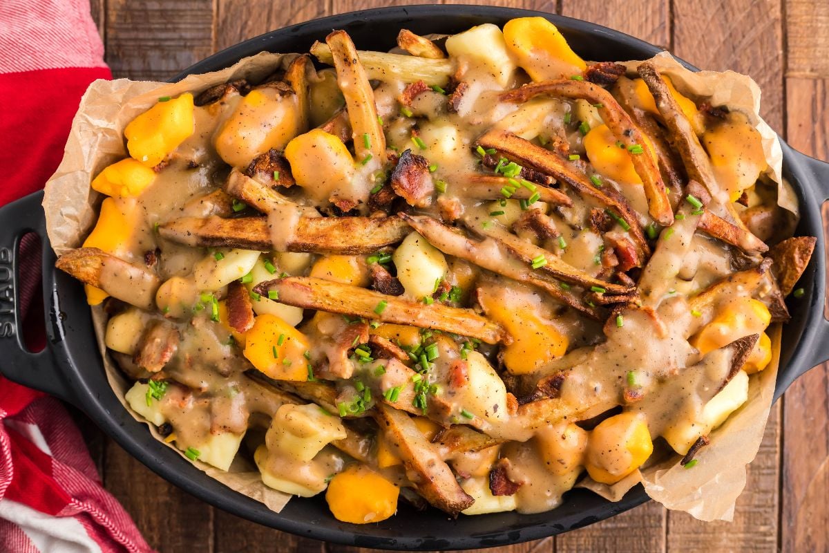 Canadian poutine with fries, cheese curds, hearty gravy, and bacon served in a black dish on a wooden table—a delicious twist on the classic poutine recipe with meat.