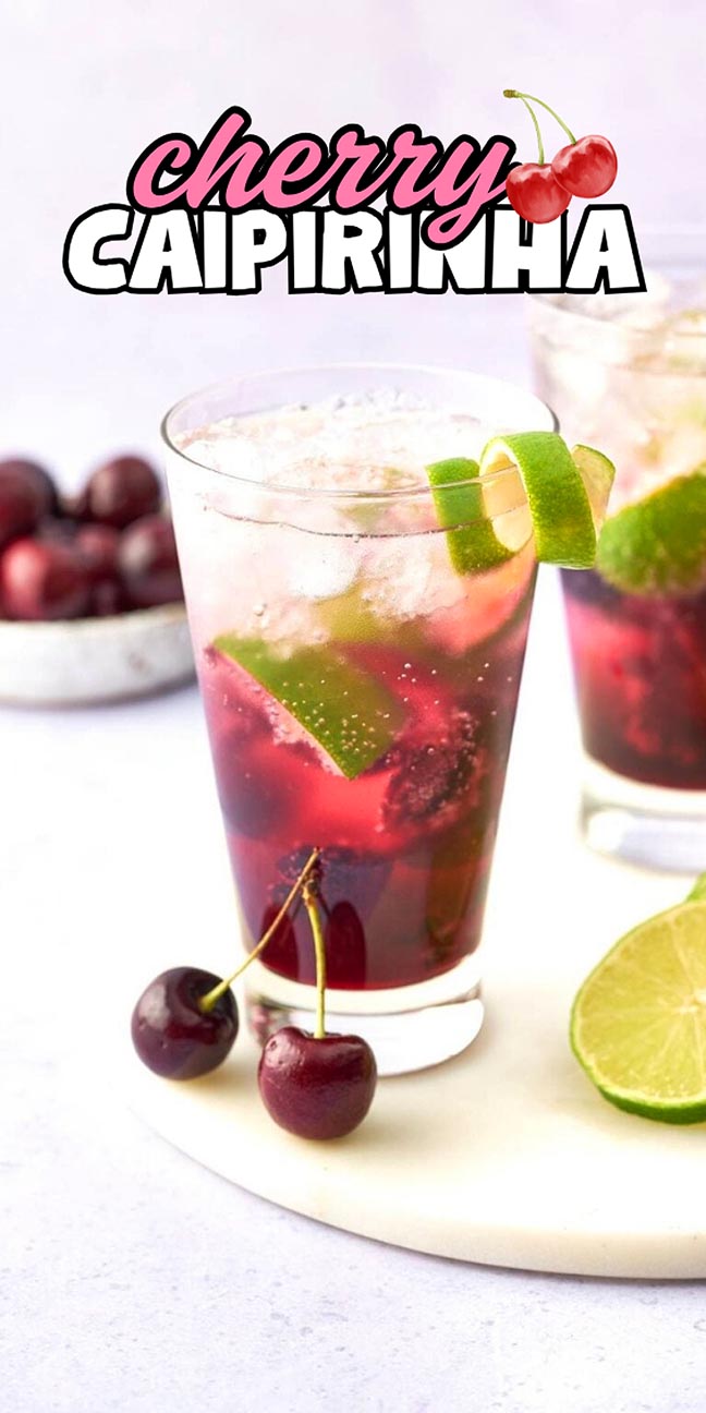 A glass of cherry caipirinha with lime slices, ice, fresh cherries, and a lime half on a white surface.