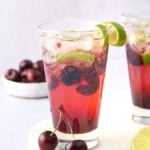 Two glasses of Cherry Caipirinha, a vibrant Brazilian cocktail, with ice, lime slices, and cherries, plus extra limes and cherries nearby—the best summer cocktail recipe for any gathering.