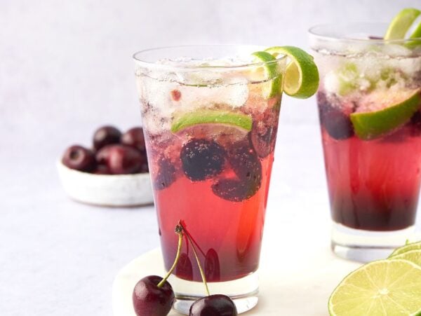 Two glasses of Cherry Caipirinha, a vibrant Brazilian cocktail, with ice, lime slices, and cherries, plus extra limes and cherries nearby—the best summer cocktail recipe for any gathering.