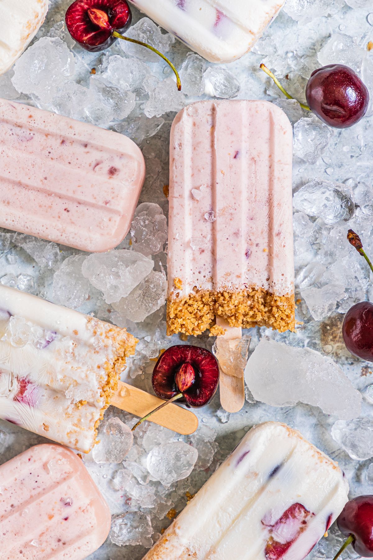 Assorted Cherry Cheesecake Popsicles with crumbs on ice, surrounded by fresh cherries—a creamy popsicle recipe that’s perfect for summer days.