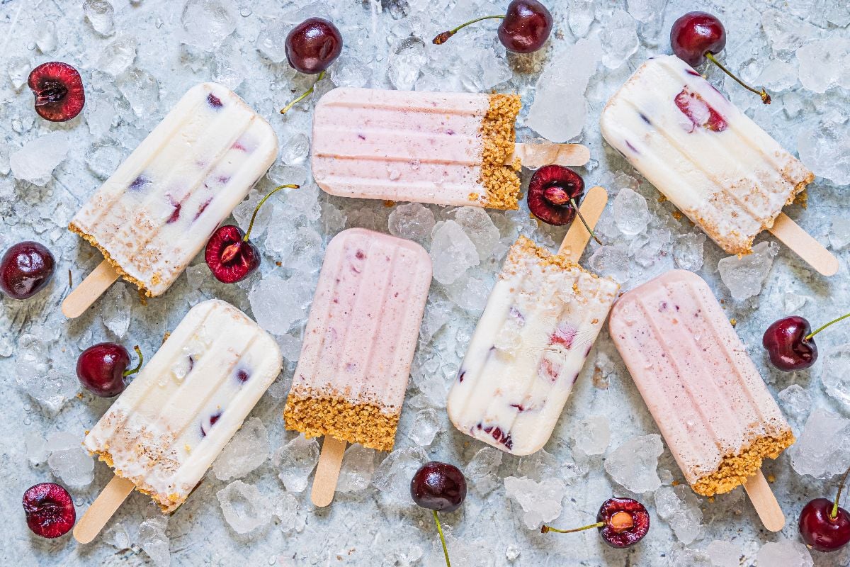 Cherry Cheesecake Popsicles with a graham cracker crust on ice, surrounded by fresh cherries—a perfect cherry dessert popsicle and summer treat recipe!.