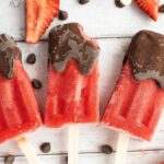 Three Chocolate Covered Strawberry Popsicles with a rich chocolate coating, surrounded by strawberries and chocolate chips on a wooden surface—easy homemade popsicles perfect for any chocolate lover.
