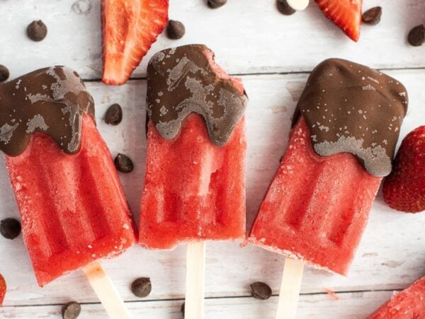 Three Chocolate Covered Strawberry Popsicles with a rich chocolate coating, surrounded by strawberries and chocolate chips on a wooden surface—easy homemade popsicles perfect for any chocolate lover.