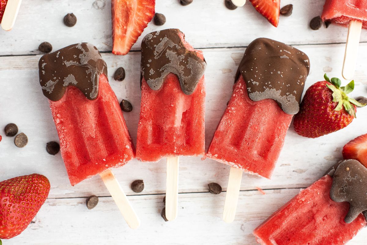 Strawberry popsicles dipped in chocolate, surrounded by fresh strawberries and chocolate chips—an easy homemade popsicles recipe perfect for a summer treat.