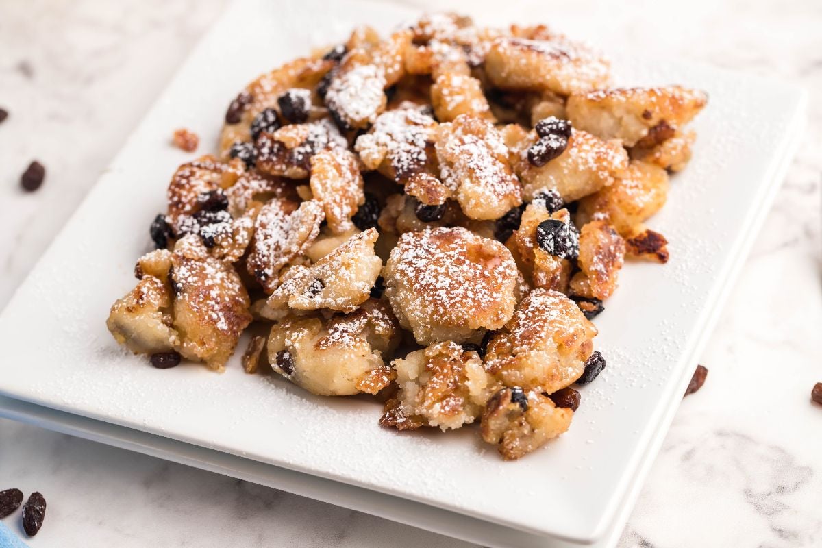 A plate of powdered sugar-dusted, scrambled Austrian and German dessert pancakes with raisins sits on a white square plate.
