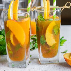 Two glasses of Hard Peach Iced Tea with peach slices, mint, and straws, surrounded by fresh peaches—perfect for a refreshing boozy iced tea recipe.