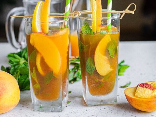 Two glasses of Hard Peach Iced Tea with peach slices, mint, and straws, surrounded by fresh peaches—perfect for a refreshing boozy iced tea recipe.