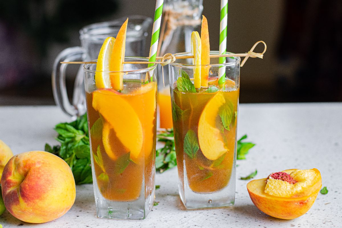 Two tall glasses of Hard Peach Iced Tea with peach slices, mint, and striped straws, surrounded by fresh peaches.