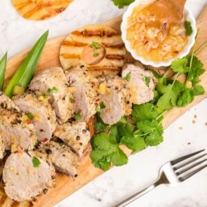 Sliced grilled pork with pineapple, cilantro, and sauce on a wooden board with a fork on the side—an easy Instant Pot Hawaiian pork tenderloin for a delicious Hawaiian inspired recipe.