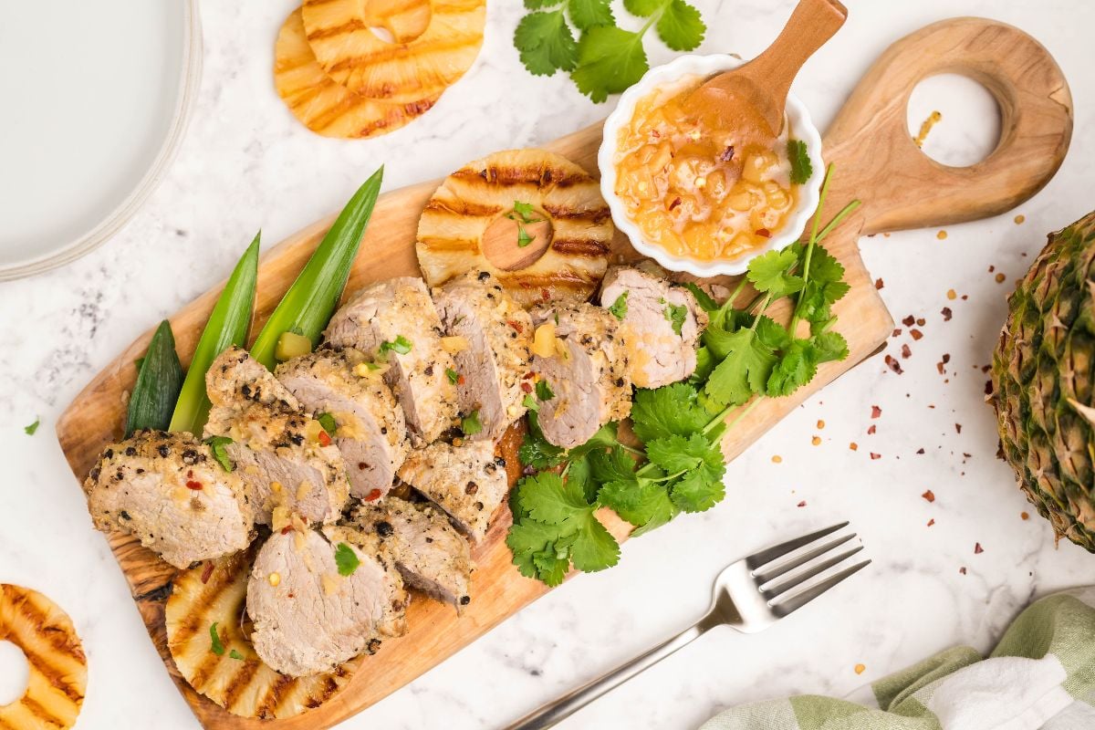 Sliced Instant Pot Hawaiian pork tenderloin with pineapple, fresh cilantro, okra, and sauce on a wooden board, on a marble surface.