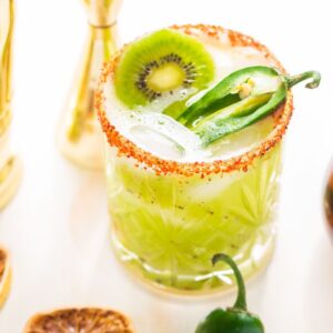 A zesty Kiwi Jalapeño Margarita features a green cocktail with kiwi and jalapeño slices, a chili salt rim, and ice, served beside a syrup bottle and dried citrus.
