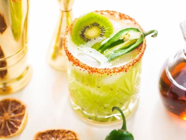 A zesty Kiwi Jalapeño Margarita features a green cocktail with kiwi and jalapeño slices, a chili salt rim, and ice, served beside a syrup bottle and dried citrus.