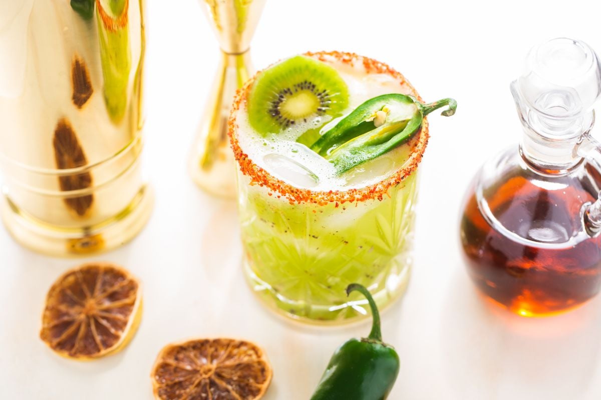 A green Kiwi Jalapeño Margarita with ice, kiwi, and jalapeño slices, rimmed with chili powder, sits beside dried citrus and syrup—a bold cocktail using tequila and orange liqueur for the best spicy margarita experience.