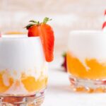 Two glasses of creamy orange and white frozen mango cocktail, garnished with strawberries and red-striped straws—a vibrant layered slushie drink recipe perfect for summer.