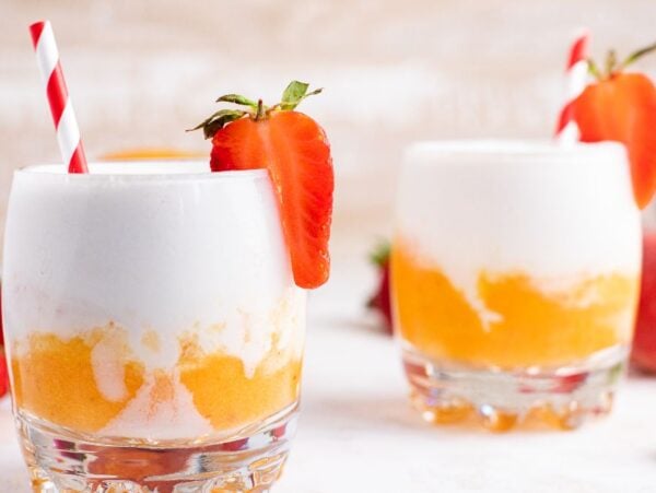 Two glasses of creamy orange and white frozen mango cocktail, garnished with strawberries and red-striped straws—a vibrant layered slushie drink recipe perfect for summer.