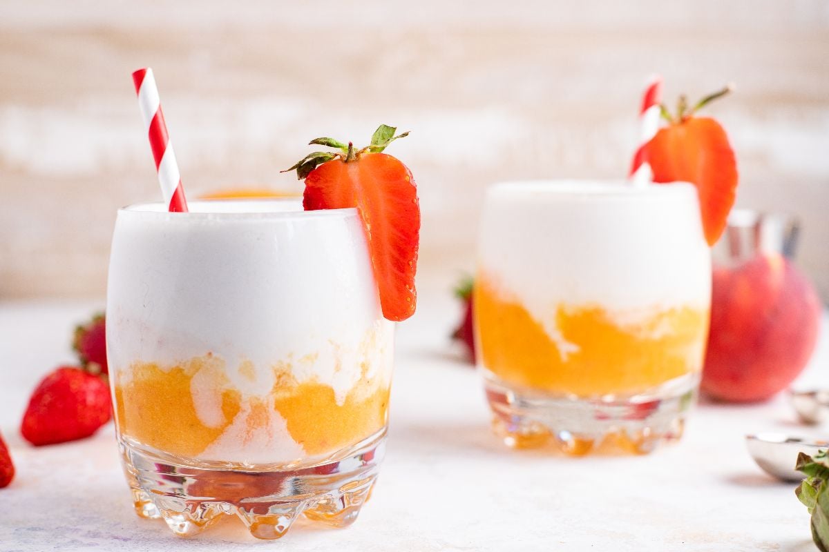 Two glasses of layered mango colada slushie with vibrant yellow and white layers, each topped with a strawberry and striped straw, create the perfect tropical rum cocktail for summer sipping.