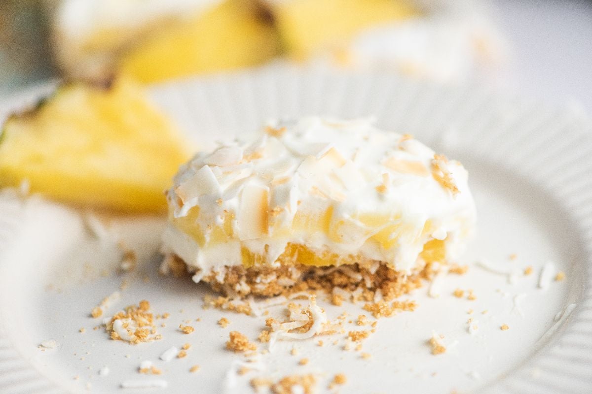 A creamy summer dessert with a crumbly crust on a white plate, featuring flavors inspired by Hawaiian pineapple and coconut cake, with a pineapple slice in the background.
