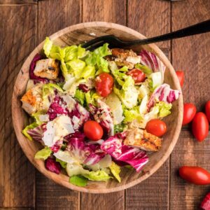 A wooden bowl of summer BBQ chicken salad with cherry tomatoes, croutons, and greens sits on a wooden table with a black fork.