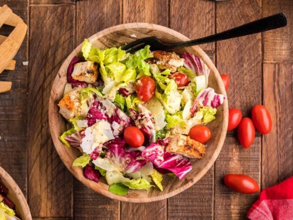 A wooden bowl of summer BBQ chicken salad with cherry tomatoes, croutons, and greens sits on a wooden table with a black fork.