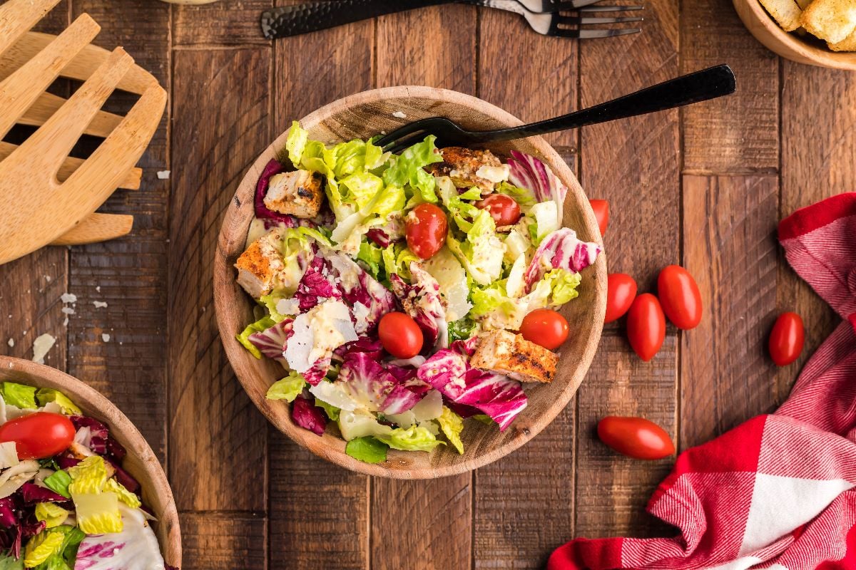 Wooden bowl of simple radicchio salad with lettuce, tomatoes, croutons, and cheese on a wooden table, served with a fork and napkin.