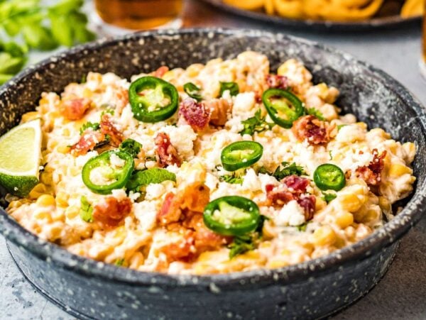 A bowl of creamy jalapeño corn dip topped with bacon, cheese, and cilantro, served with lime wedges on the side—a delicious slow cooker dip recipe perfect for any gathering.