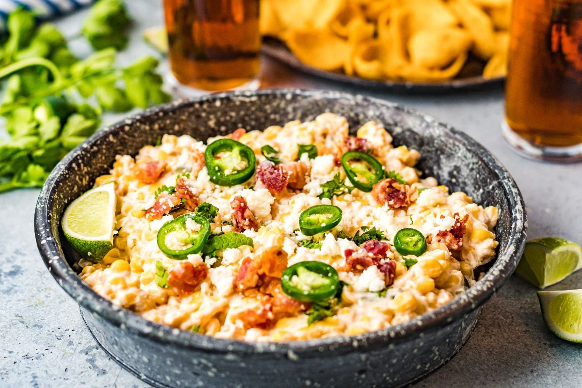 A bowl of cheesy Mexican-style appetizer, this creamy corn dip is topped with sliced jalapeños, bacon, cheese, and served with lime wedges—a perfect Slow Cooker Jalapeño Corn Dip for your next gathering.
