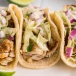 Three fish tacos topped with shredded cabbage, diced red onion, and creamy sauce, served with lime wedges and a zesty garlic cilantro lime coleslaw on the side.