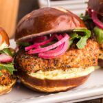 Three spicy chicken sandwiches on pretzel buns with arugula, pickled onions, and creamy wasabi mayo sauce on a plate.