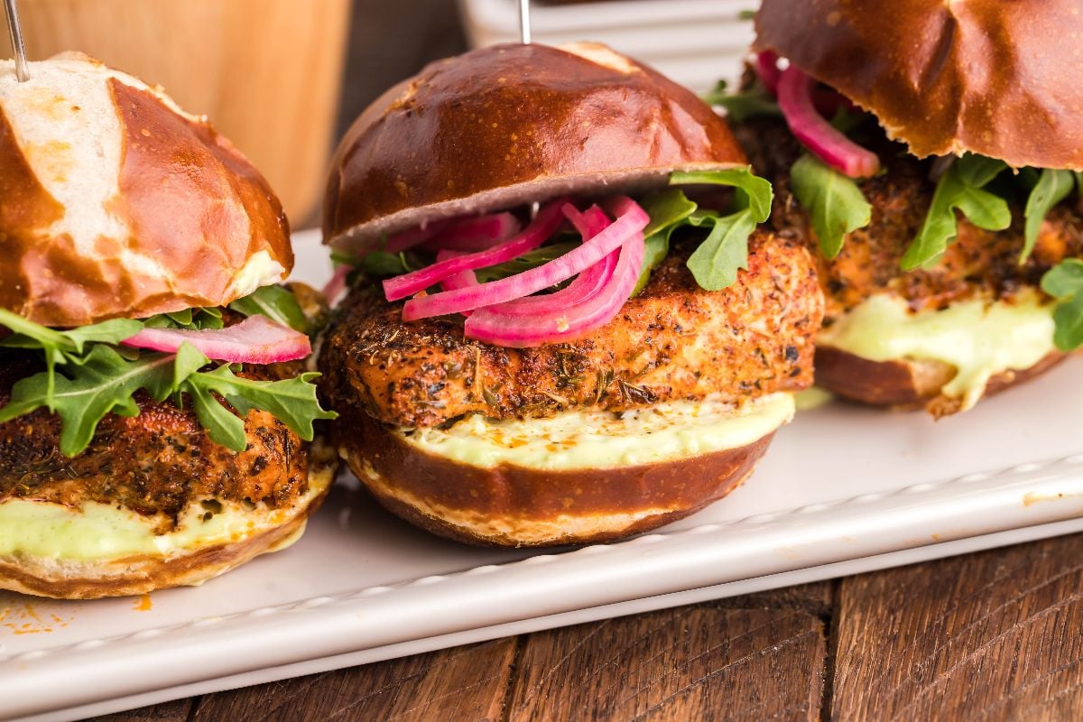 Three chicken sliders on pretzel buns with arugula, pickled onions, and Wasabi Mayo are served on a white platter—perfect as a zesty starter for your next Summer BBQ salmon burgers or smoked salmon recipe feast.