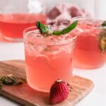 Two glasses of pink iced drinks with basil and strawberries on a wooden board—a refreshing Strawberry gin rickey made with strawberry basil simple syrup. A pitcher and towel sit in the background.