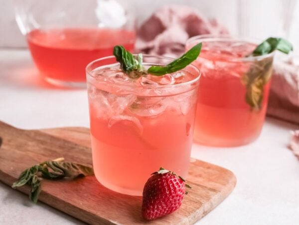Two glasses of pink iced drinks with basil and strawberries on a wooden board—a refreshing Strawberry gin rickey made with strawberry basil simple syrup. A pitcher and towel sit in the background.