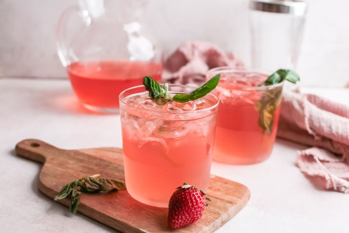 Two pink drinks with ice, basil, and a strawberry sit on a wooden board, with a pitcher in the background—a refreshing summer cocktail and a vibrant variation on a classic gin rickey.