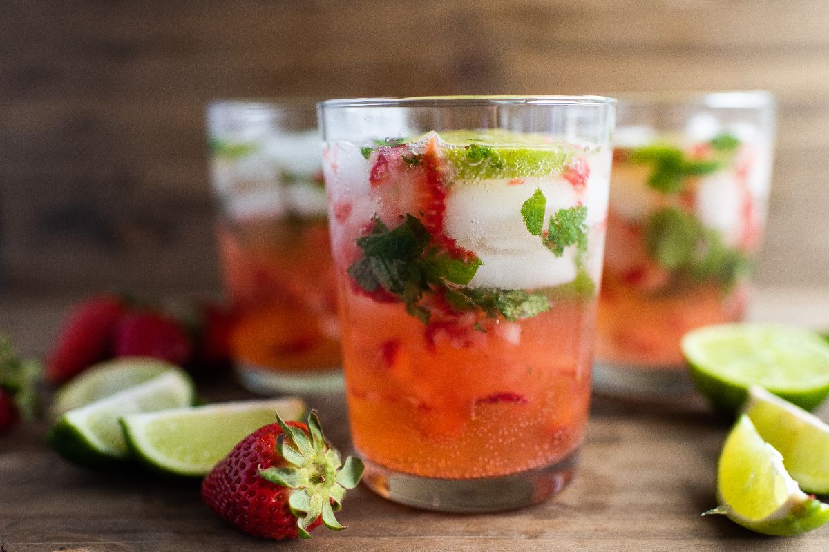 A glass of strawberry and lime cocktail with ice, mint, and fresh fruit slices on a wooden surface—a refreshing drink with strawberries and your choice of cachaça or dark rum.
