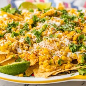 Plate of Street Corn Nachos topped with corn, cheese, cilantro, spices, and lime wedges for a unique nacho topping twist.