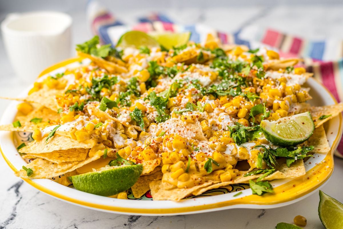 A plate of Street Corn Nachos topped with corn, crema, cheese, cilantro, chili powder, and lime wedges—bringing you unique nacho toppings inspired by classic elote and nacho chips.