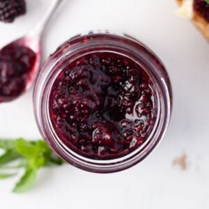 A jar of sugar free blackberry jam with a spoonful of jam and fresh blackberries beside it on a white surface.