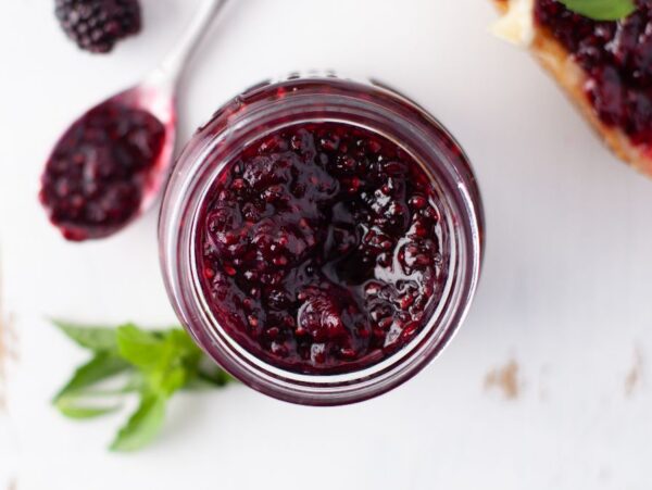 A jar of sugar free blackberry jam with a spoonful of jam and fresh blackberries beside it on a white surface.
