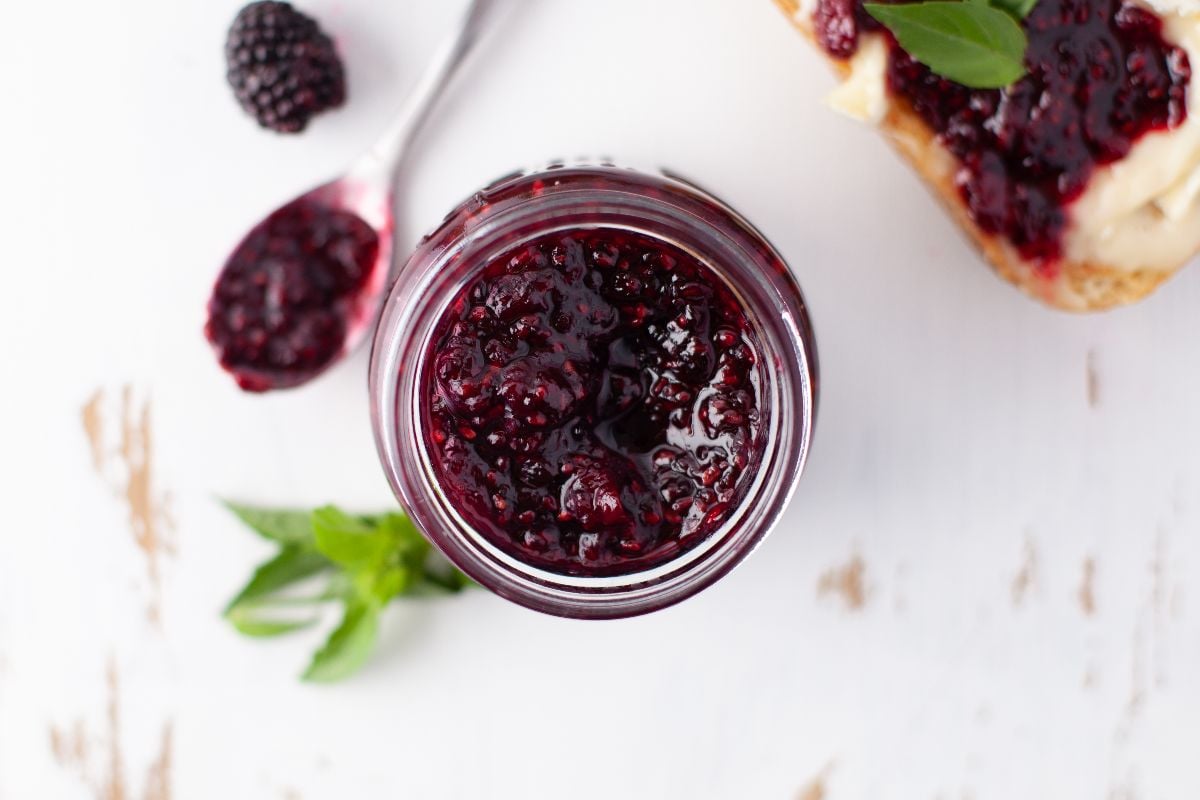 Top view of a jar of blackberry jam with a spoon, fresh berries, and mint leaves on a white surface—perfect for the easiest jam recipe or a healthy homemade jam recipe using your favorite natural sweeteners.