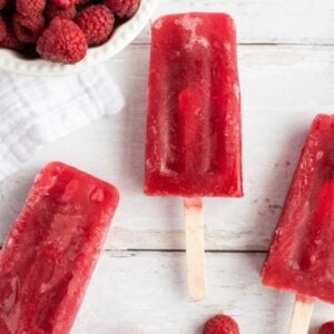 Four raspberry popsicles and loose raspberries on a white surface, with a bowl of raspberries nearby—these popsicles are made with sweetened ice tea, using frozen or fresh raspberries for a refreshing treat.