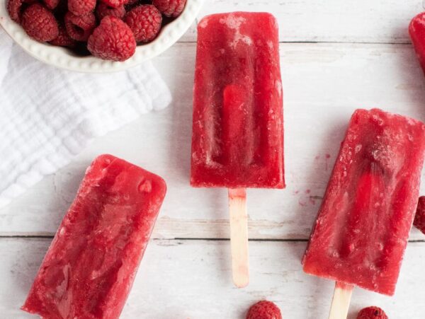 Four raspberry popsicles and loose raspberries on a white surface, with a bowl of raspberries nearby—these popsicles are made with sweetened ice tea, using frozen or fresh raspberries for a refreshing treat.
