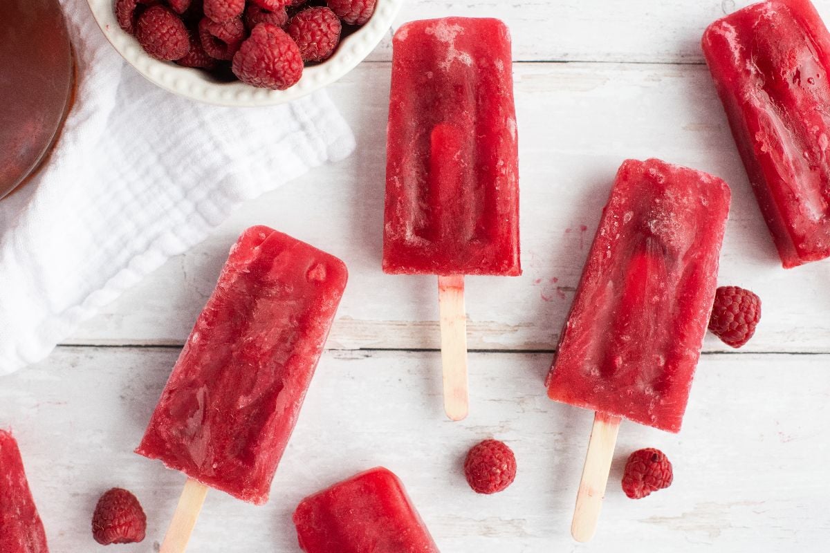 Five red raspberry popsicles made using frozen or fresh raspberries sit beside fresh berries on a white wooden surface with a white towel nearby—perfect sweet summer treats.