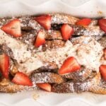 A baked French toast casserole topped with powdered sugar, strawberries, and a dollop of cream in a white baking dish—perfect for an overnight French toast breakfast.